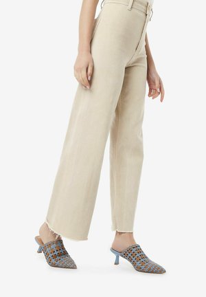 Jeans beige a vita alta con gamba larga abbinati a mule con tacco blu e marroni, con apertura sul retro e punta affusolata.