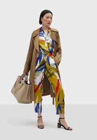 Trench coat beige sopra una tuta multicolore con motivi astratti in giallo, blu e rosso. Borsa beige e sandali neri con tacco.