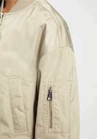 Beige Bomberjacke mit geripptem Kragen und silbernem Reißverschlussfach am Oberarm, auf der linken Körperseite getragen, vor einem schlichten Hintergrund.