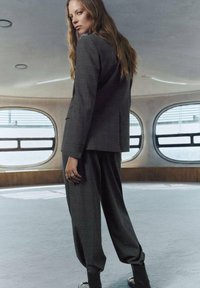 Traje a medida de cuadros grises con una chaqueta ajustada y pantalones holgados. El modelo tiene el pelo largo y lleva un anillo y botas negras hasta el tobillo. El fondo tiene ventanas curvas.