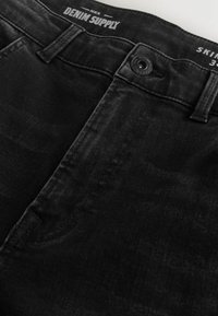 Jeans skinny negri din denim cu o textura netedă, având un buton superior și un design cu cinci buzunare. Numele brandului este vizibil pe banda de la talie.