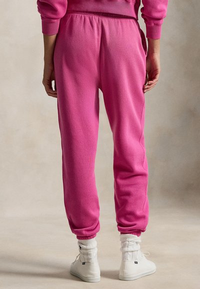 Ροζ φόρμα με ελαστική μέση και μανσέτες, υφή φλις, κωνικά πόδια, συνδυασμένη με λευκά ψηλοτάκουνα παπούτσια και πλεκτές κάλτσες.