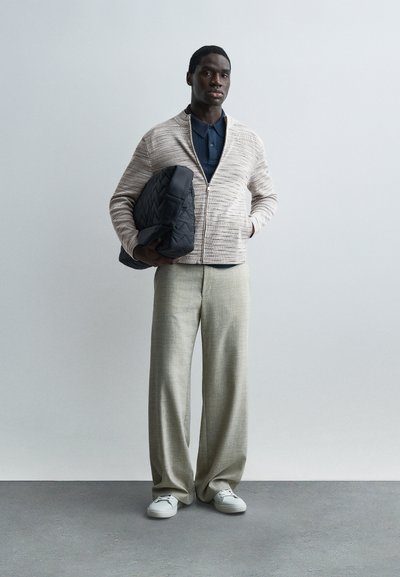 Homme portant une veste beige zippée, un polo bleu marine, un pantalon large beige, des baskets blanches, tenant un sac matelassé noir sur un fond gris uni.