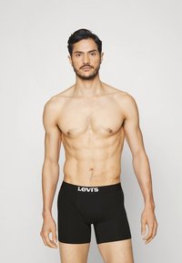 Črni bokserji z elastičnim pasom, ki imajo logotip Levi's, gladko teksturo, oprijeto zasnovo in dolžino do sredine stegna. Model prikazuje sprednji pogled.