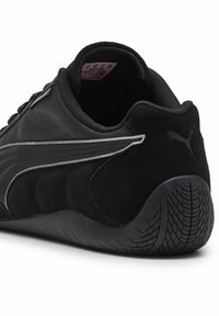 Zapatilla deportiva negra con materiales de cuero liso y gamuza, que presenta una franja curva plateada y una suela de goma texturizada.