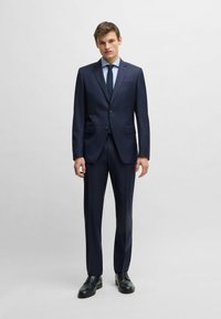 Granatowy garnitur z marynarką na dwa guziki, spodniami z dżerseju oraz jasnoniebieską koszulą w kratkę. Stylizację dopełniają wypolerowane czarne skórzane buty.