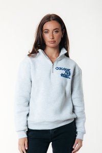 Mujer con cabello largo castaño que lleva un sudadera de media cremallera gris claro con texto azul "Colourful Rebel", pantalones negros y un collar dorado.