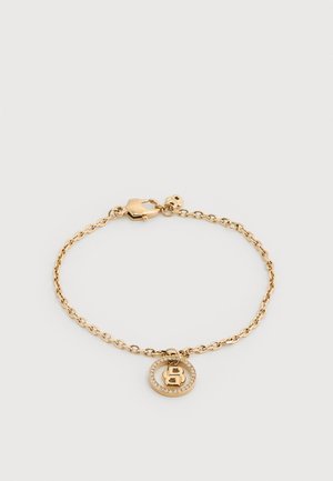 Zelta ķēdes aproce ar apaļu piekariņu, kuram ir rhinestone apmale un centrā izcelts burts "B", fiksēta ar omāra aizdari.