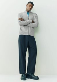 Grau zipfeliger Pullover, darunter ein blaues Hemd, lockere schwarze Hose und schwarze Schuhe, mit glatten Texturen und kontrastierenden Farben.