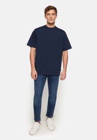 Un tricou din bumbac bleumarin, cu guler rotund și mâneci scurte, asortat cu blugi slim-fit și adidași albi, pe un fundal simplu.