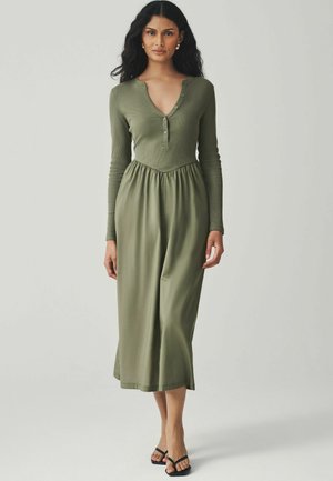 Olivgrünes, langärmeliges Kleid mit Henley-Ausschnitt, elastischer Taille und fließendem Rock, der bis zur Mitte der Wade reicht; weiche, gerippte Textur. Schwarze Sandalen.