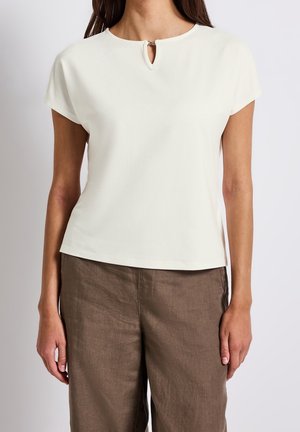 Vrouw draagt een wit shirt met korte mouwen en een klein gaatje aan de voorkant, gecombineerd met loszittende bruine broek, tegen een effen achtergrond.