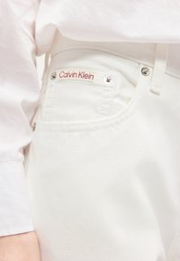 Κοντινό πλάνο τσέπης λευκού τζιν με το κόκκινο λογότυπο Calvin Klein σε μικρή ετικέτα, φορεμένη με λευκό πουκάμισο.