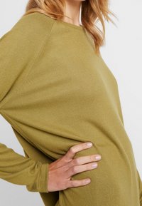 Olive green long-sleeve top with a relaxed fit, ribbed texture, wide neckline, and raglan sleeves, highlighting side seam and fabric drape.