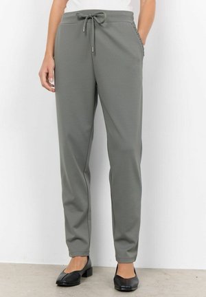 Woman wearing gray drawstring pants with side pockets and black low-heeled shoes, standing against a white wall.