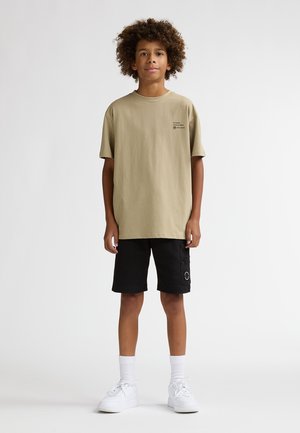 Tricou bej din bumbac cu decolteu rotund, asortat cu pantaloni scurți negri cargo cu buzunare laterale. Look-ul este completat de adidași albi și șosete până la gleznă.