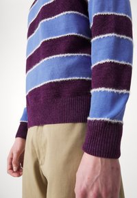 Personne portant un pull rayé violet et bleu avec un pantalon beige clair, debout, les mains détendues le long du corps.