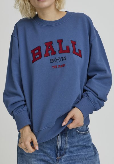 Sweat-shirt bleu avec texte brodé rouge "BALL 1974 THE JEANS" sur le devant, doté de poignets et d'un ourlet côtelés, porté sur un jean en denim clair.
