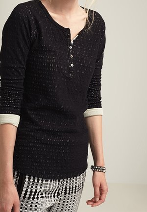 Persoon draagt een zwart getextureerd langemouwshirt met witte manchetten en zwart-wit geblokte broek, toont een armband met studs aan de linkerpols.