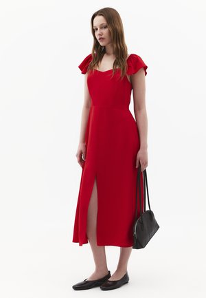 Femme portant une robe midi rouge avec des manches volantées et une fente frontale, tenant un sac à main noir, debout sur fond blanc.
