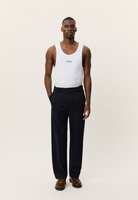 Άσπρη βαμβακερή φανέλα χωρίς μανίκια με στρογγυλή λαιμόκοψη και μαύρο έμβλημα, συνδυασμένη με ναυτικό παντελόνι κομμένο στα μέτρα και καφέ παπούτσια. Απλός και κλασικός σχεδιασμός.