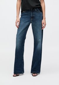 Παντελόνια τζιν σκούρου μπλε με φαρδύ πόδι, κατασκευασμένα από ντένιμ, με κλασικό σχεδιασμό πέντε τσεπών και ελαφρώς ξεθωριασμένη υφή. Συνδυασμένα με μαύρο τοπ.