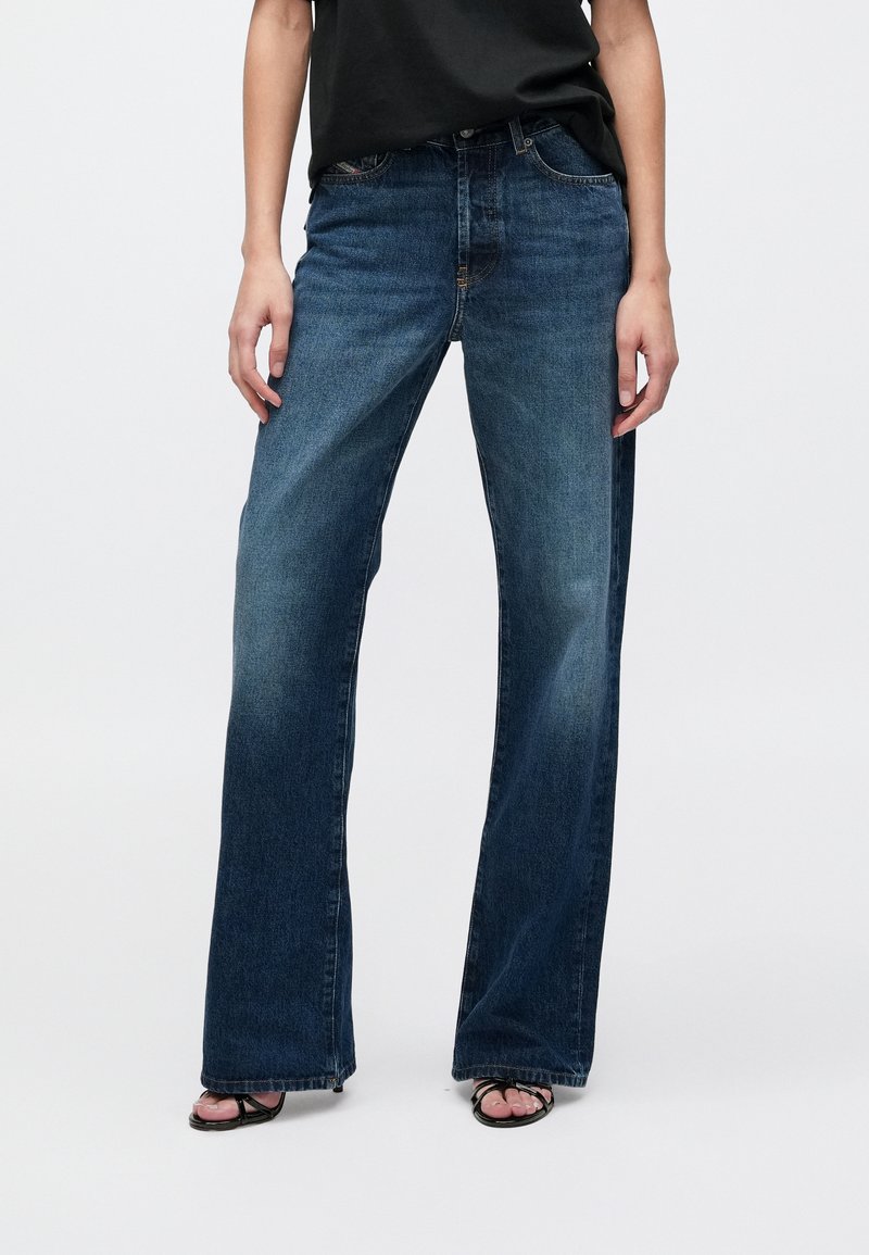 Παντελόνια τζιν σκούρου μπλε με φαρδύ πόδι, κατασκευασμένα από ντένιμ, με κλασικό σχεδιασμό πέντε τσεπών και ελαφρώς ξεθωριασμένη υφή. Συνδυασμένα με μαύρο τοπ.