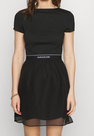 Black dress with short sleeves; fitted bodice and flowing skirt. Features a Calvin Klein Jeans logo waistband and a sheer hemline.