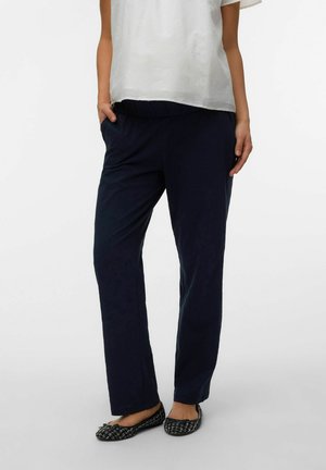 Naine, kes kannab musti sirge säärega pükse küljetaskutega, valget lühikeste varrukatega pluusi ja musti mustrilisi madalaid jalanõusid.