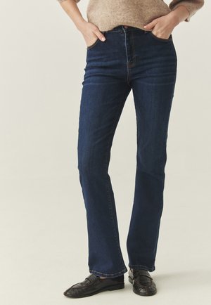 Person iført mørkeblå jeans med lige ben, beige strikket sweater og sorte læder loafers, stående med hænderne i lommerne.