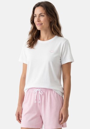 Kobieta o falowanych brązowych włosach, ubrana w biały t-shirt z małą różową buźką oraz jasnoróżowe szorty ze sznurkiem, lekko się uśmiechająca.