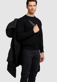 Maglione nero a maglia e jeans scuri indossati da un modello che tiene un piumino nero sulla spalla, mostrando un design elegante e minimalista.