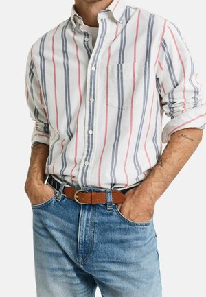 Mężczyzna ubrany w białą koszulę na guziki z czerwonymi i niebieskimi pionowymi paskami, niebieskie dżinsy i brązowy skórzany pasek, ręce w kieszeniach.