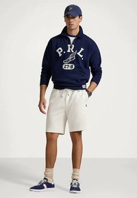 Ναυτικό πουλόβερ με λευκή γραφή, λευκά σορτς με κορδόνια και μπλε αθλητικά παπούτσια. Χακί κάλτσες μέχρι τον αστράγαλο. Ναυτικός σκούφος στο κεφάλι.