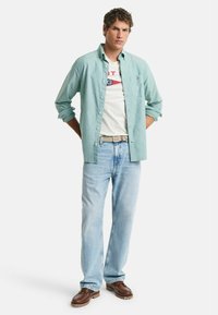 Lysegrøn skjorte med knapper over en hvid t-shirt med et logo, sammen med lyseblå jeans og brune sko. Modellen står med hænderne bag ryggen.