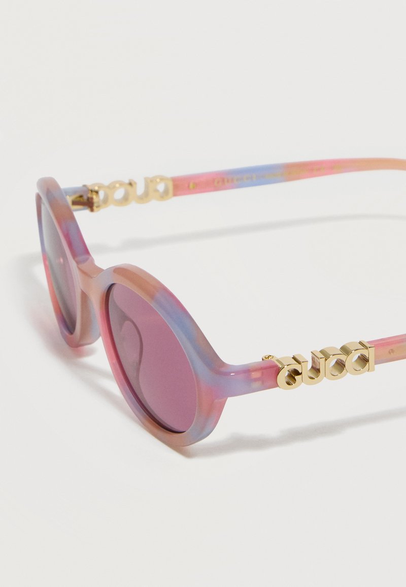 Lunettes de soleil rondes avec montures translucides roses et bleues, verres violets et logo Gucci en or sur les branches.