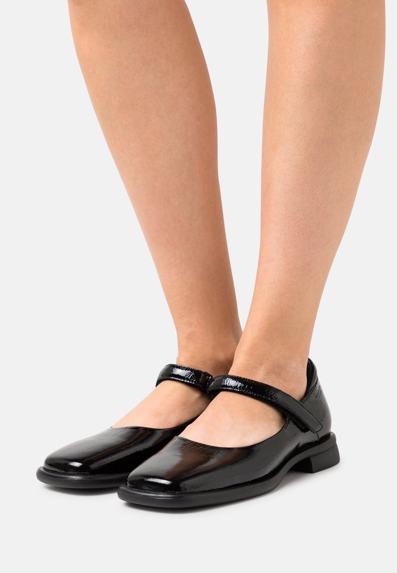Chaussures Mary Jane noires brillantes avec talons bas et une bride unique sur le dessus, portées sur des jambes nues devant un fond uni.