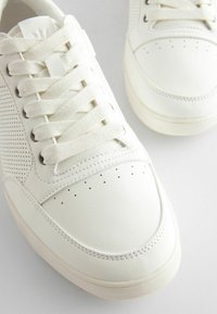 Białe sneakersy z perforowanymi elementami, płaskimi sznurówkami oraz gładką skórzaną fakturą, z zaokrąglonym noskiem i gumową podeszwą.