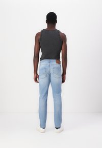 Jeans de mezclilla azul claro con ajuste slim y bolsillos traseros; llevados con una camiseta de tirantes oscura y zapatillas blancas, vista desde atrás.