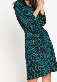 Svart klänning med turkosa diamanter, lång ärm och ett midjeband. Mjuk, texturerad tyg, knälång design med en avslappnad silhuett.