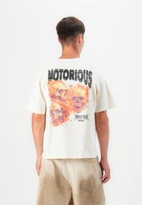 Cream-colored cotton t-shirt featuring a graphic of three skulls surrounded by orange flames, with "NOTORIOUS" and "WORLD TOUR 1990" text.