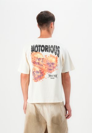 Cream-colored cotton t-shirt featuring a graphic of three skulls surrounded by orange flames, with "NOTORIOUS" and "WORLD TOUR 1990" text.