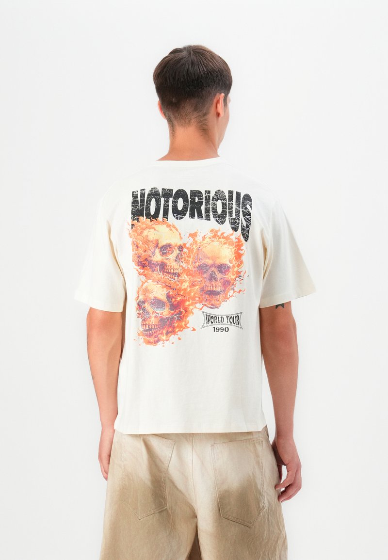 Krem majica od pamuka s grafikom od tri lubanje okružene narančastim plamenovima, s natpisima "NOTORIOUS" i "WORLD TOUR 1990".