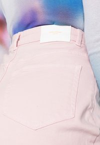 Svetlo rožnati jeans kratke hlače s prilegajočim dizajnom, ki imajo zadnje zaklopne žepe in belo oznako z napisom "VERO MODA" v oranžni barvi.