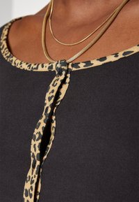 Svart topp med leopardtryck vid halsringningen, med en knutdetalj i mitten. Slät tygkvalitet med kontrasterande texturerad kant.