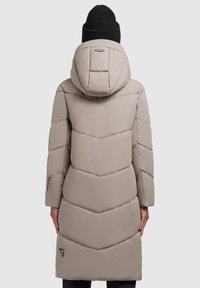 Beige padded coat with a chevron quilted design, featuring a hood and a zippered side pocket, worn with a black knit beanie.
