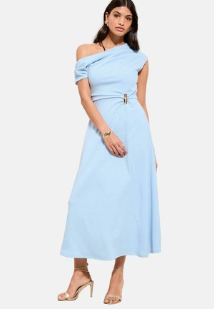 Žena v svetlomodrých midi šatách s jedným ramienkom a opaskom so zlatou prackou, v kombinácii so zlatými sandálmi na vysokom opätku a zlatými šperkmi, pózujúca na bielom pozadí.