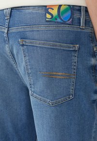 Des jeans en denim bleu présentent une poche arrière avec un détail de couture horizontal et une étiquette à rayures de couleur avec "S.Q." à la taille.