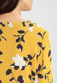 Tissu jaune avec un motif floral présentant des feuilles noires et des fleurs blanches. Le design a une texture lisse et une coupe décontractée.