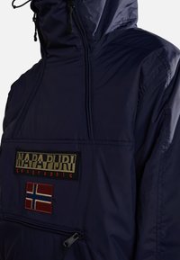 Marinblå vattentät jacka med dragkedja framtill, med en stor logotyp och en norsk flagga. Slät textur, justerbar huva.